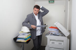 Picture of a man holding a sheet of paper next to a printer, looking confused